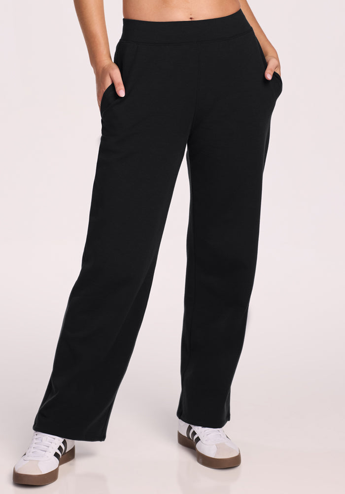 A person stands against a light background wearing Woolx Brynn Pants in black—heavyweight, loose-fitting lounge pants with hands in pockets—paired with white sneakers with brown soles; their upper body is not visible. 