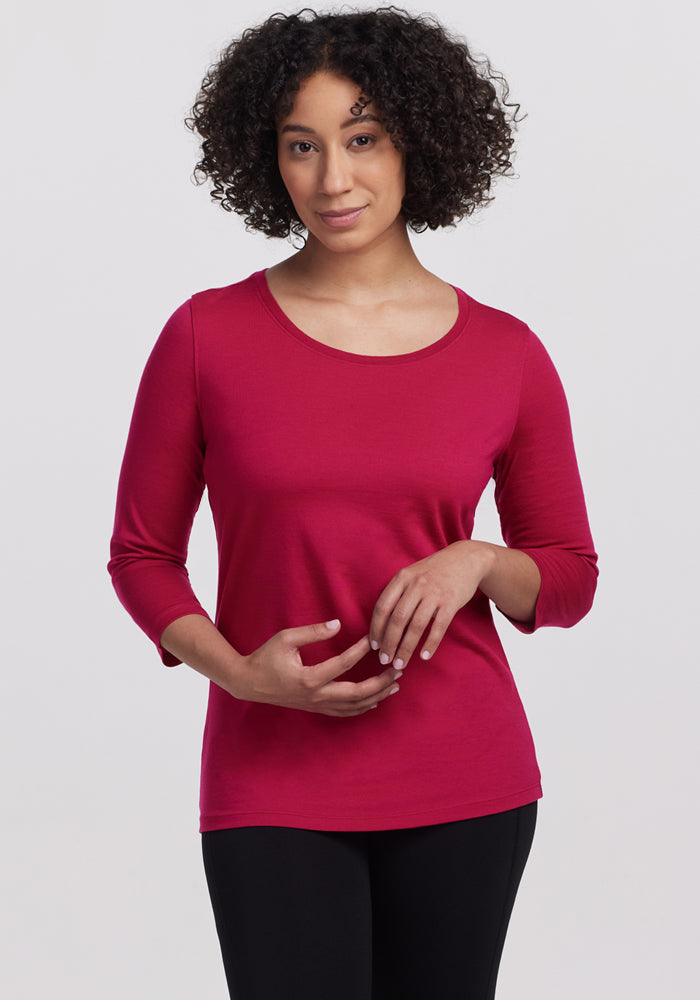 A woman with curly hair wears the Woolx Jenny 3/4 Sleeve Crew Neck in Poppy Spark and black pants. She stands against a light gray background, looking at the camera with a slight smile and her hands relaxed in front of her. 