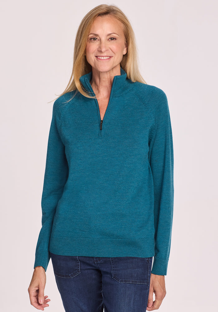 A woman with long blonde hair wears the Woolx Collins Half Zip Sweater in Harbor Teal and dark jeans, standing and smiling against a plain light background. 