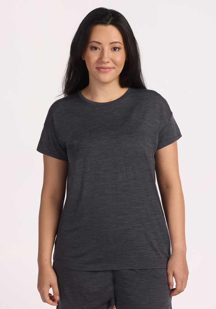 A woman with long dark hair smiles gently at the camera, wearing the Woolx Billie Lounge Tee in Cinder Melange and matching pants; both are moisture-wicking and she stands against a plain light background. 