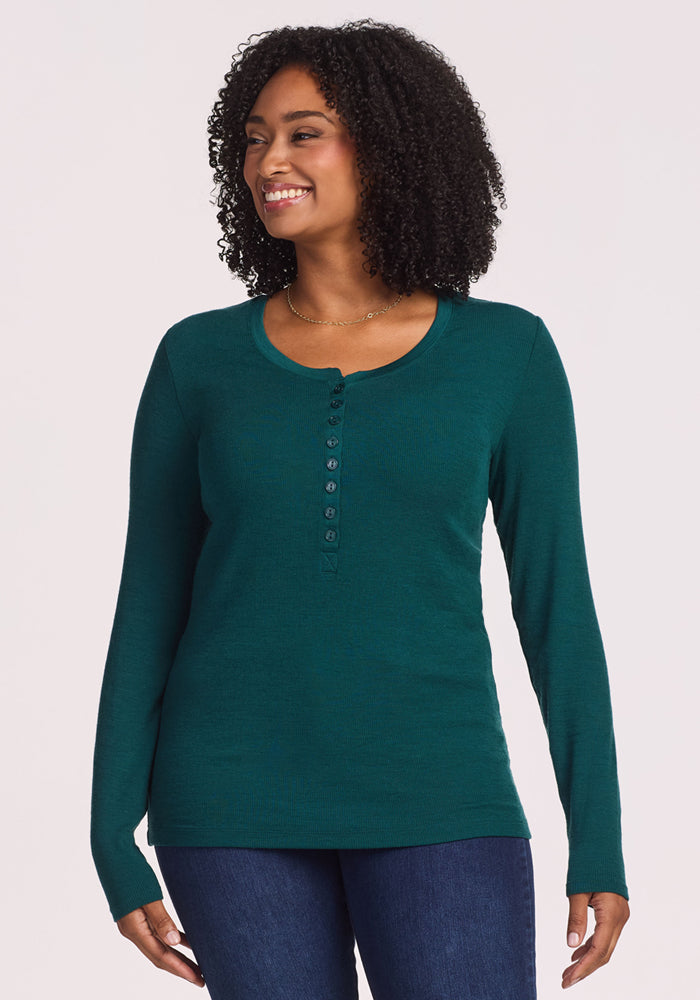 A woman with curly hair smiles while wearing the Woolx Reese Ribbed Henley in Riverbank Green and blue jeans, standing against a plain, light background. 
