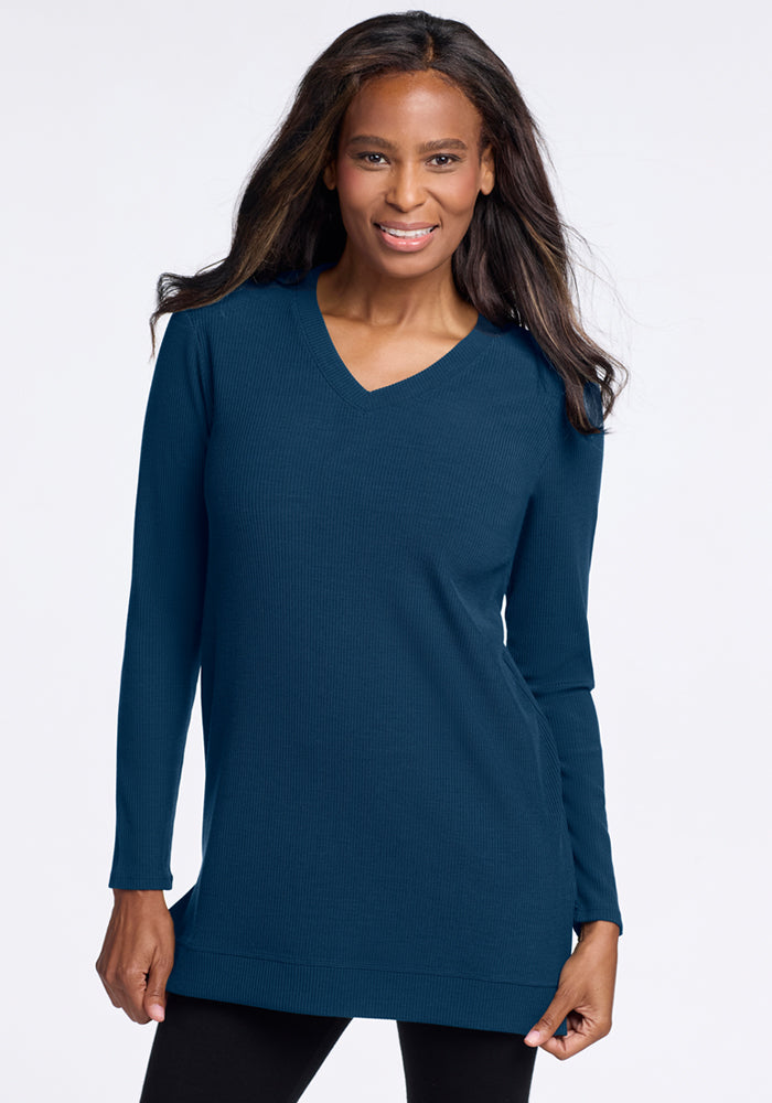 A woman with long dark hair smiles while wearing the Woolx Evie Ribbed Tunic in Majolica Blue with a V-neck and black pants, standing against a plain light background—perfect for temperature regulating comfort. 
