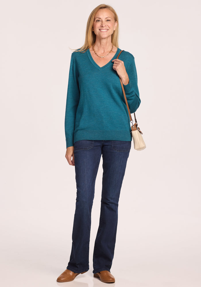 A woman smiles against a light background, wearing the Woolx Francesca Sweater in Harbor Teal, paired with dark blue jeans, brown shoes, and a beige crossbody bag.