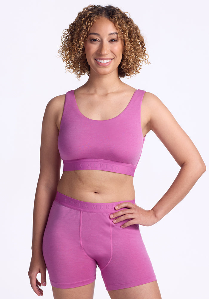A woman with curly hair smiles at the camera, wearing the Woolx Aubrey Lounge Bra in Wild Blossom with a matching logo band and boxer shorts, set against a plain white background.