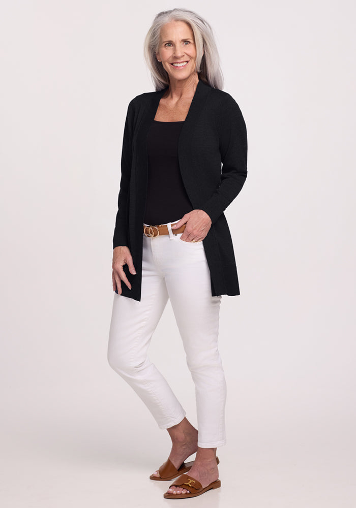 A smiling older woman with long gray hair wears the Woolx Macie Sweater in black over a matching top, paired with white pants and brown accessories, standing against a plain light background.