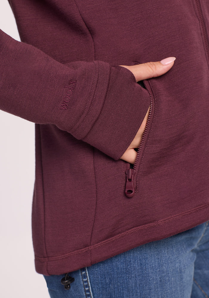 A person wears the Woolx Cubby Hooded Sweatshirt in Grape Harvest, featuring thumbholes, zippered side pockets, and "WOOLX" embroidered near the wrist, paired with blue jeans.