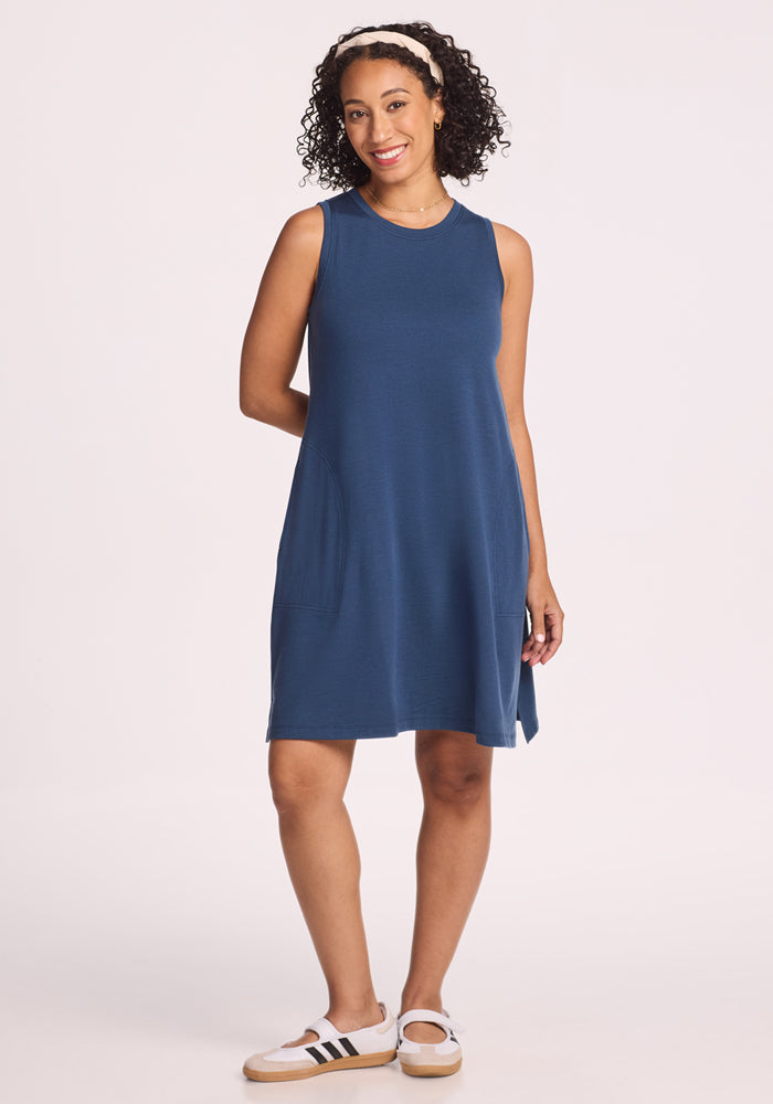 A woman with curly hair smiles against a plain light background, wearing the Woolx Clara Dress - Starry Night, a sleeveless, knee-length blue summer dress with pockets, paired with white sneakers featuring black stripes. 