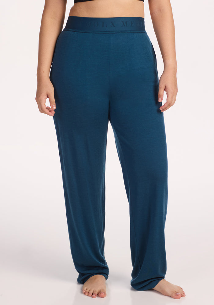 A person stands barefoot in Woolx Maya Lounge Pants – Majolica Blue, featuring an elastic waistband. The loose-fitting Merino wool pants are shown from midriff to feet, set against a plain white background. 