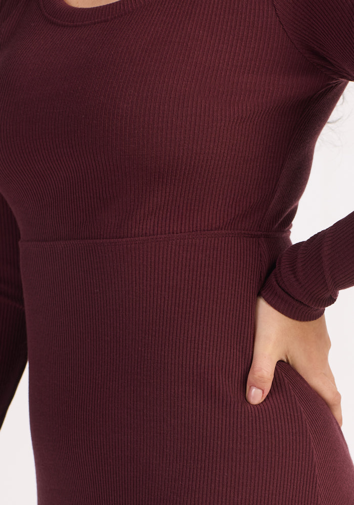 A close-up shows a person in the Woolx Caroline Ribbed Twirl Dress in Toasted Garnet. The ribbed merino style features long sleeves, with one hand on their hip. Only the torso and upper arm appear against a plain background.