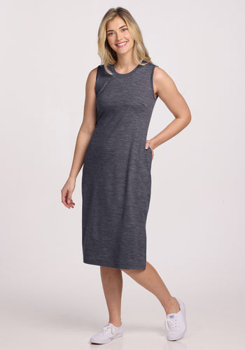 A woman with blonde hair smiles with one hand in her pocket, wearing the Woolx Cassie Dress in Cinder Melange—a sleeveless, knee-length, wrinkle-resistant Merino wool piece—paired with white sneakers against a plain white background. | Ariana is 5'11", wearing a size S