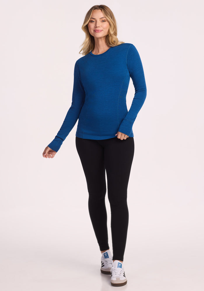 A woman with blonde hair smiles softly, standing against a plain background in the Woolx Hannah Long Sleeve Top - Nightfall, paired with black leggings and white sneakers. One hand is slightly raised.