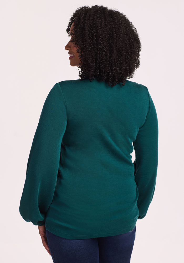 A woman with curly hair is shown from the back, wearing the Woolx Charlotte Puff Sleeve Top in Riverbank Green and dark blue jeans, standing against a plain light background.
