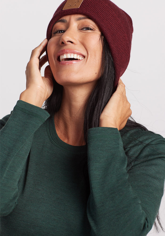 A smiling woman in the Woolx Blair Tunic - Forest and a maroon knit beanie poses with her hands on her hat, looking upward against a light background, enjoying lightweight warmth and odor-resistant comfort.