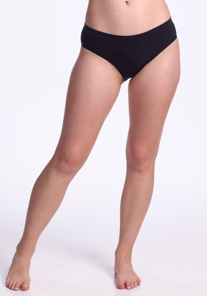 A person stands barefoot against a white background, wearing the Woolx Maeve Bikini in black. The image is cropped from mid-torso down, highlighting the lower body and legs.