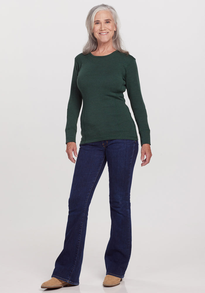 A smiling older woman with long gray hair stands against a white background, wearing the Woolx Hannah Long Sleeve Top in Forest, paired with blue jeans and tan shoes.