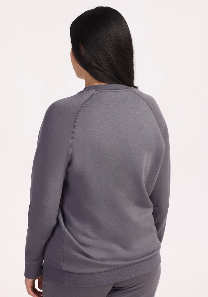 A woman with long dark hair is shown from the back, wearing the Woolx Bailey Crewneck Sweatshirt in Arctic Dusk and matching pants, standing against a plain light background.
