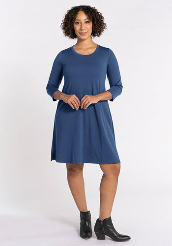 A woman stands facing forward, wearing the Woolx Rissa A Line Dress in Starry Night—a knee-length, long-sleeve blue merino wool dress—paired with black ankle boots, hands lightly clasped, perfect for fall and winter against a plain white background. | Tori is 5'7", wearing a size S