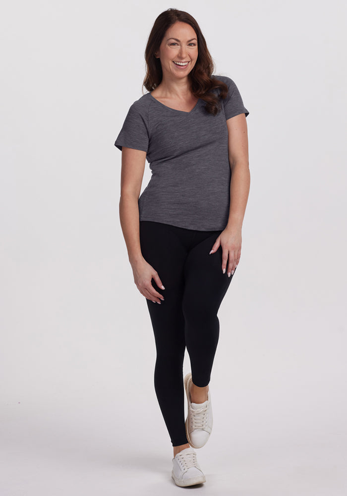 A woman with long brown hair smiles against a plain background, wearing the Woolx Mia V Neck in Cinder Melange, black leggings, and white sneakers. One leg is slightly bent and her arms rest casually at her sides.