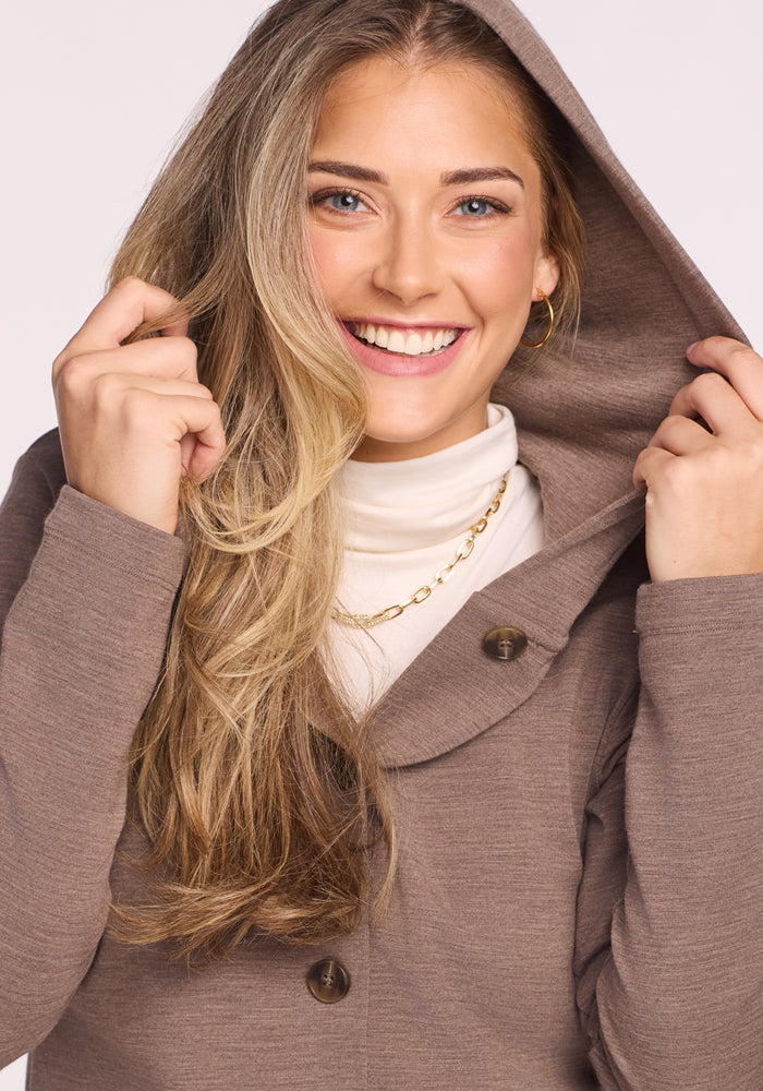 A smiling woman with long blonde hair wears the Woolx Edna Hooded Cardigan in Simply Taupe over a white turtleneck and gold chain necklace, holding the hood with both hands.