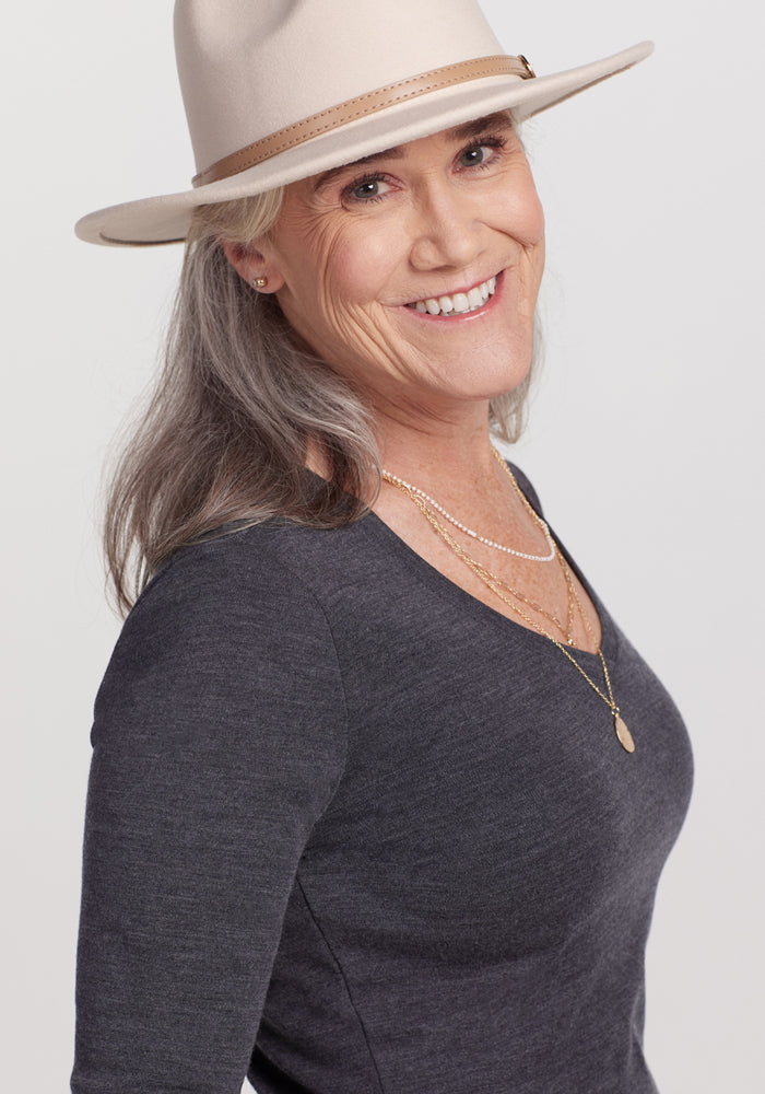 Smiling older woman with long gray hair in a light wide-brim hat, layered gold necklaces, and the Woolx Eva Tunic - Cinder Melange, posing against a plain light background—a true winter wardrobe essential.