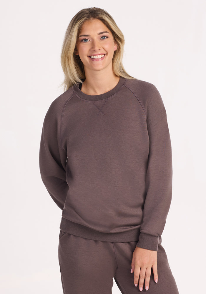 A smiling blonde woman wears the Woolx Bailey Crewneck Sweatshirt in Plum Truffle, made with a recycled polyester exterior and Merino wool interior, paired with matching brown sweatpants, standing against a plain white background. 