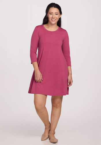 A woman smiles in a Woolx Rissa A Line Dress - Mulberry, knee-length and long-sleeved, made of Merino wool for temperature regulation and odor resistance. She has long dark hair and beige shoes against a plain light background. Perfect for any occasion!. | Sarah is 5’8”, wearing a size S