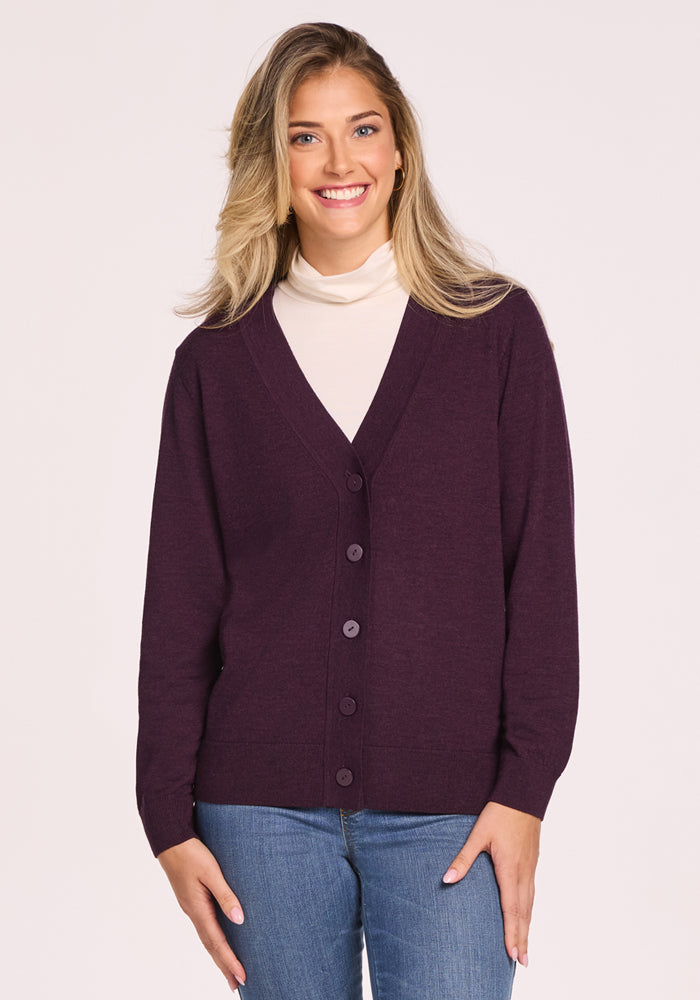 A woman with long blonde hair smiles while wearing the Woolx Gia Sweater in Deep Plum over a white turtleneck and blue jeans against a plain light background. 