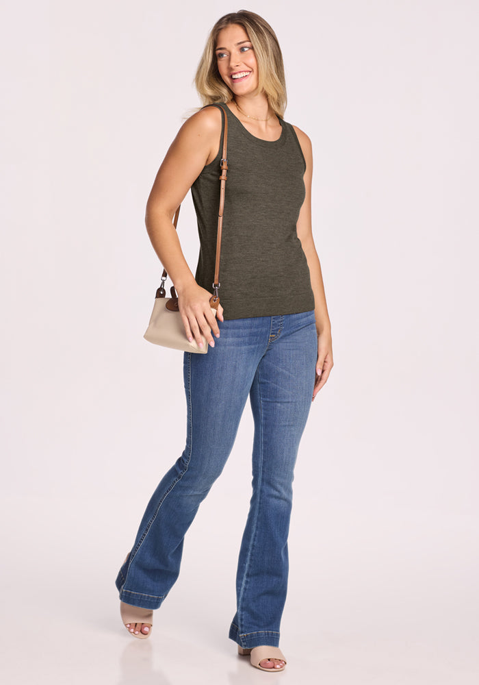 A woman wearing the Woolx Chessy Tank Top in Pine Needle, blue jeans, and beige heeled sandals stands smiling while holding a small light-colored crossbody purse against a plain white background.
