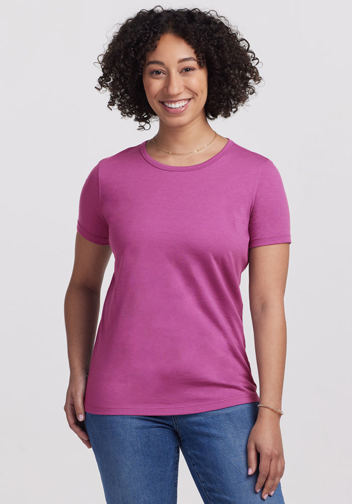 A smiling woman with curly hair wears the Woolx Liza Crew Neck Tee in Purple Wine and blue jeans, standing against a plain light background. 