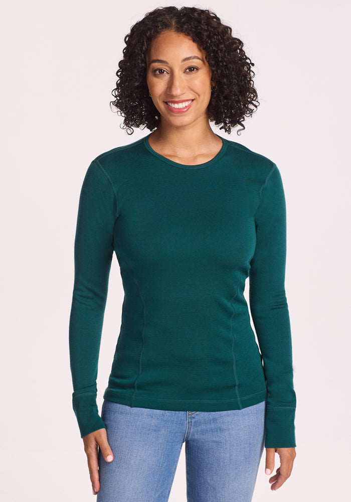 A woman with curly hair smiles at the camera, wearing the Woolx Hannah Long Sleeve Top in Riverbank Green and light blue jeans, standing against a plain white background. 