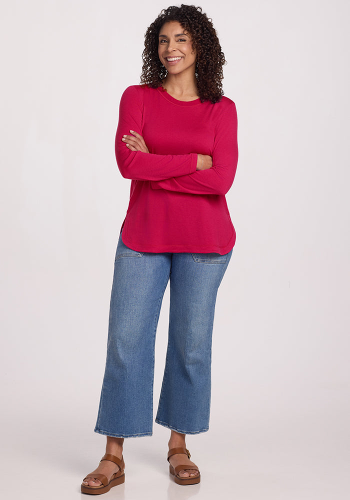 A woman with curly hair smiles, standing with arms crossed in a Woolx Hazel Tunic - Poppy Spark, blue cropped jeans, and tan sandals against a plain light background.