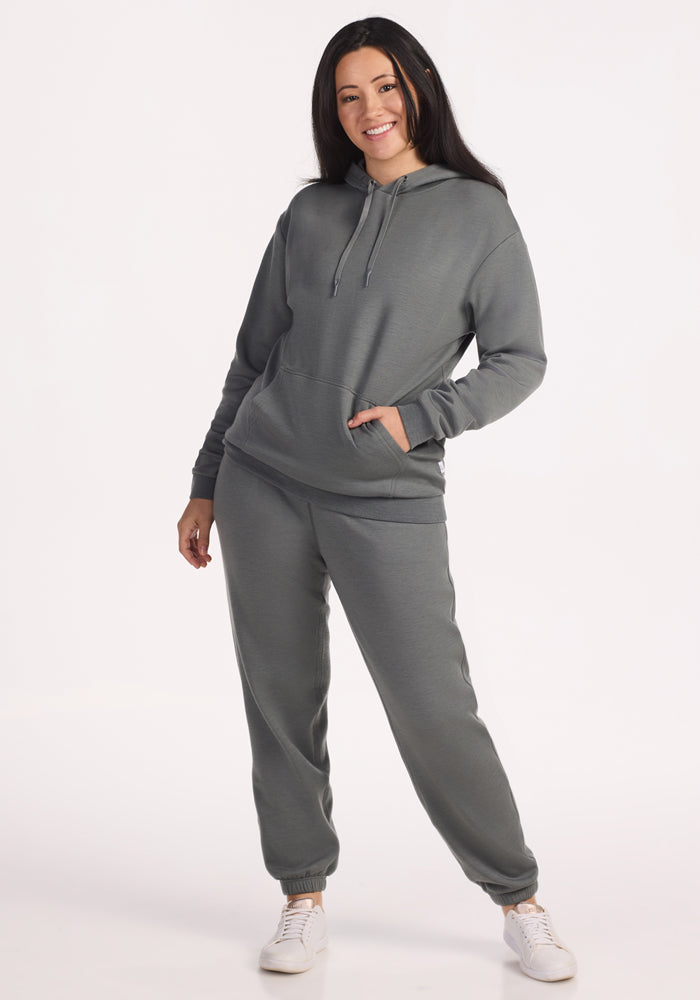 A woman with long dark hair smiles, wearing the Woolx Avery Hoodie in Sedona Sage paired with sweatpants and white sneakers. She stands against a plain white background, one hand in her pocket.