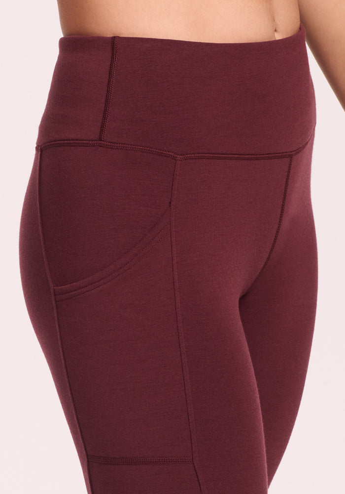Close-up of a person wearing Woolx Piper Pocket Petite leggings in Toasted Garnet, featuring visible stitching and an outer side pocket, set against a light pink background.