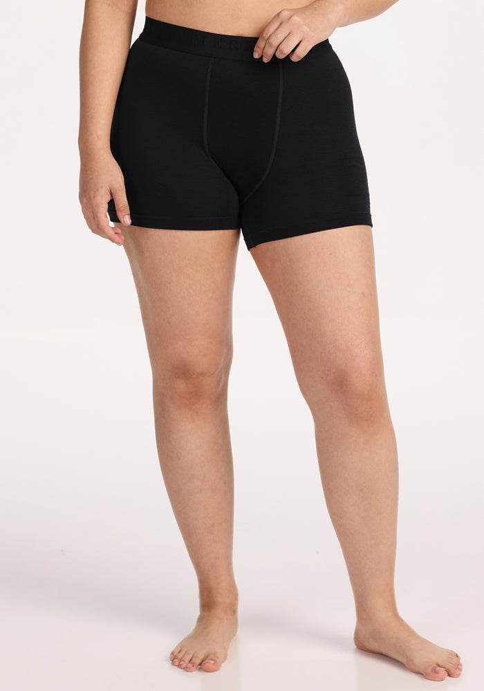 A person stands barefoot against a plain background, wearing the Woolx Elliott Boyshort in black and a matching top, with one hand on their hip. Their face is not visible. 