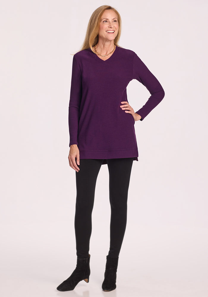A smiling woman with long blond hair wears the Woolx Evie Ribbed Tunic in Majestic Purple, paired with black leggings, boots, and a necklace. She stands hand-on-hip against a plain light background—ideal for temperature regulating comfort.