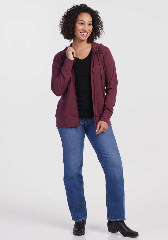 A woman with curly hair smiles while standing against a plain white background. She wears the Woolx Ryann Hoodie in Grape Harvest over a black shirt, paired with blue jeans and black ankle boots.