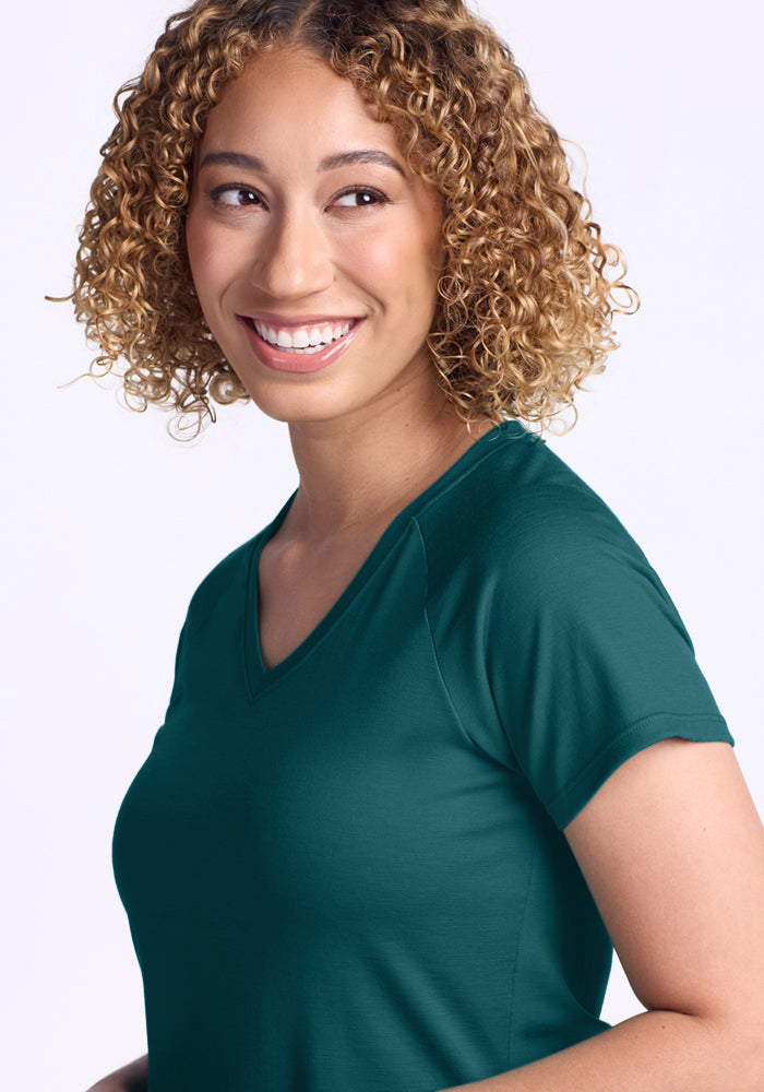 A smiling person with short, curly hair models the Woolx Mia V Neck T-shirt in Riverbank Green, standing against a plain light background and looking slightly to the side.