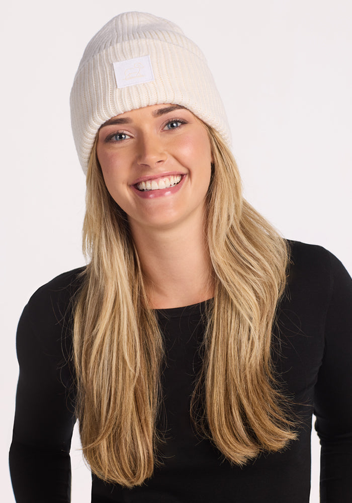 A smiling woman with long blonde hair wears the Woolx Ria Chunky Beanie in Buttercream and a black long-sleeve shirt, standing against a plain white background.