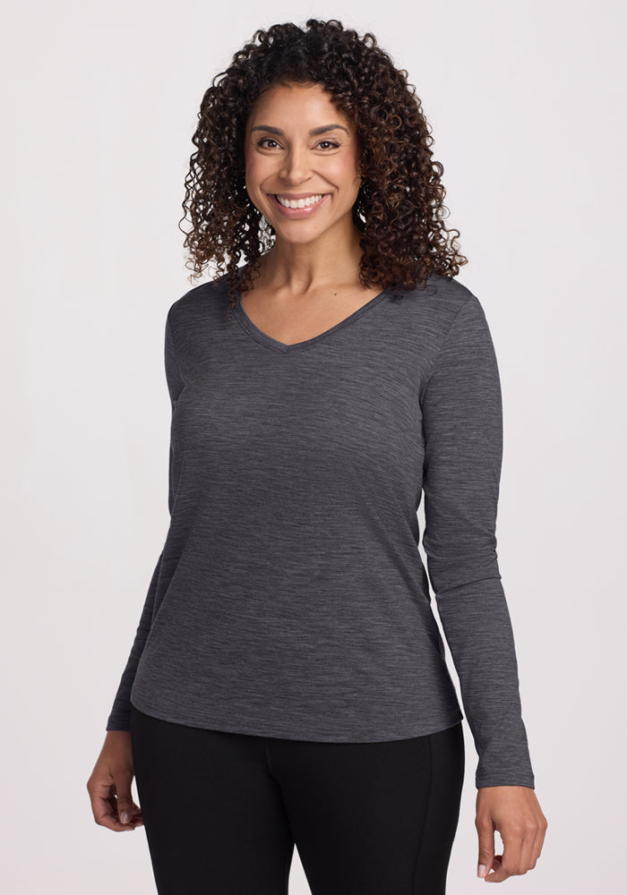 A woman with curly hair smiles in the Woolx Millie Long Sleeve - Cinder Melange and black pants, standing against a plain light background. 