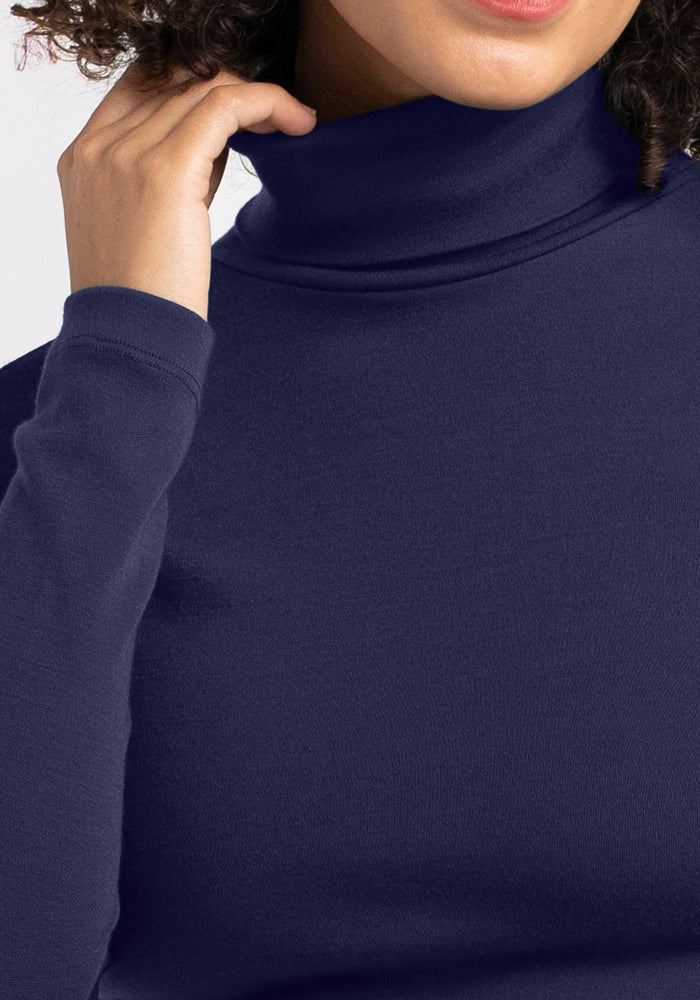 A person wearing the Woolx Peyton Turtleneck in Midnight Sapphire gently touches the collar near their face, with only the lower half of their face and upper torso visible.