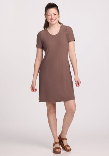 A person smiles, wearing the Woolx Harlowe Ribbed Dress - Toasted Almond, a temperature-regulating, knee-length short-sleeved dress. They pair it with brown sandals and have long hair tied back while standing on a plain white background.