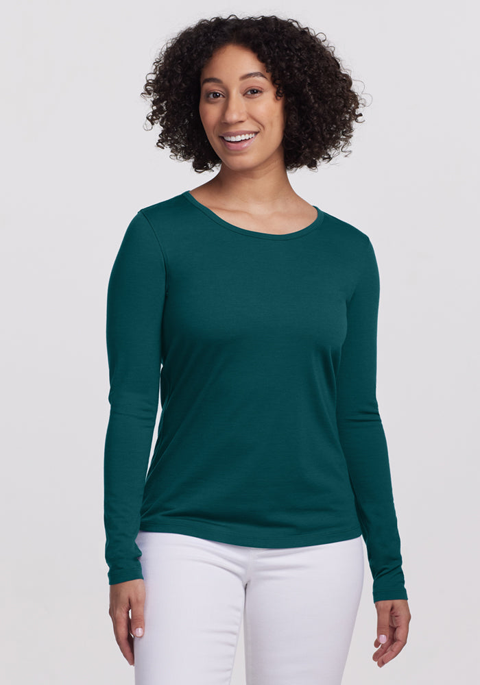 A woman with curly hair smiles in a Woolx Remi Long Sleeve T-Shirt in Riverbank Green and white pants, standing against a plain light background. 