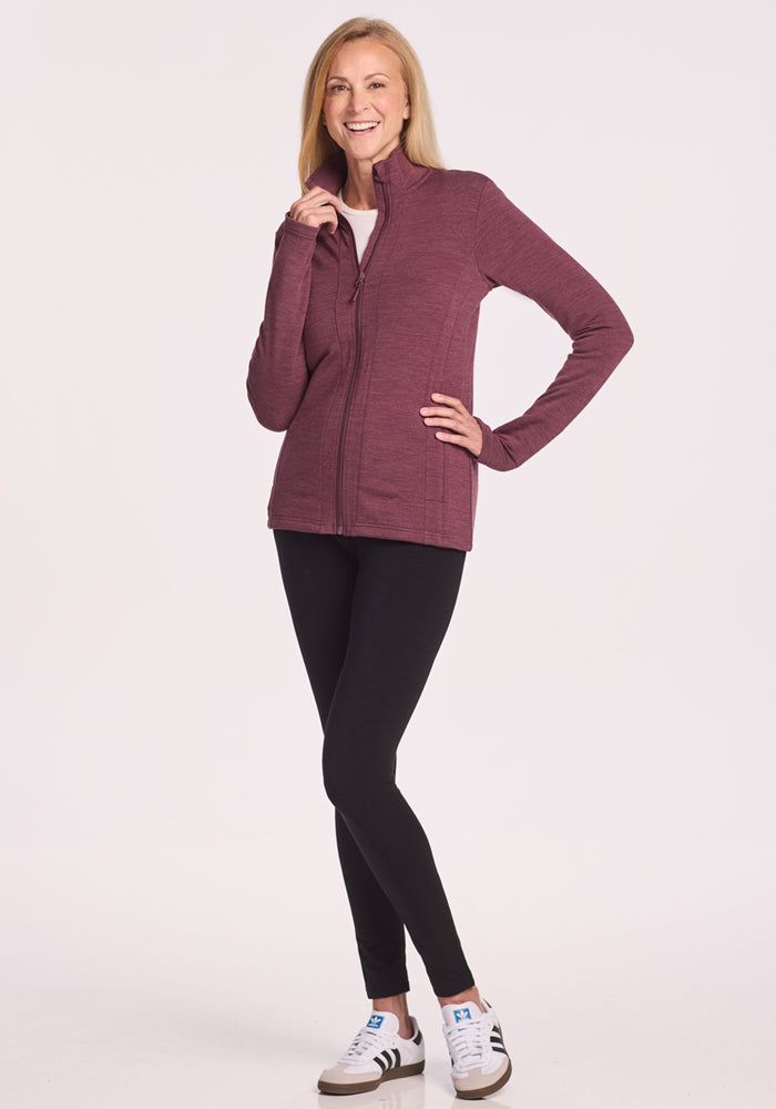 A woman smiles in the Woolx Finley Zip Up Sweatshirt in Grape Harvest Melange, paired with black leggings and white sneakers, standing against a plain white background. One hand touches her chin, the other rests on her hip.