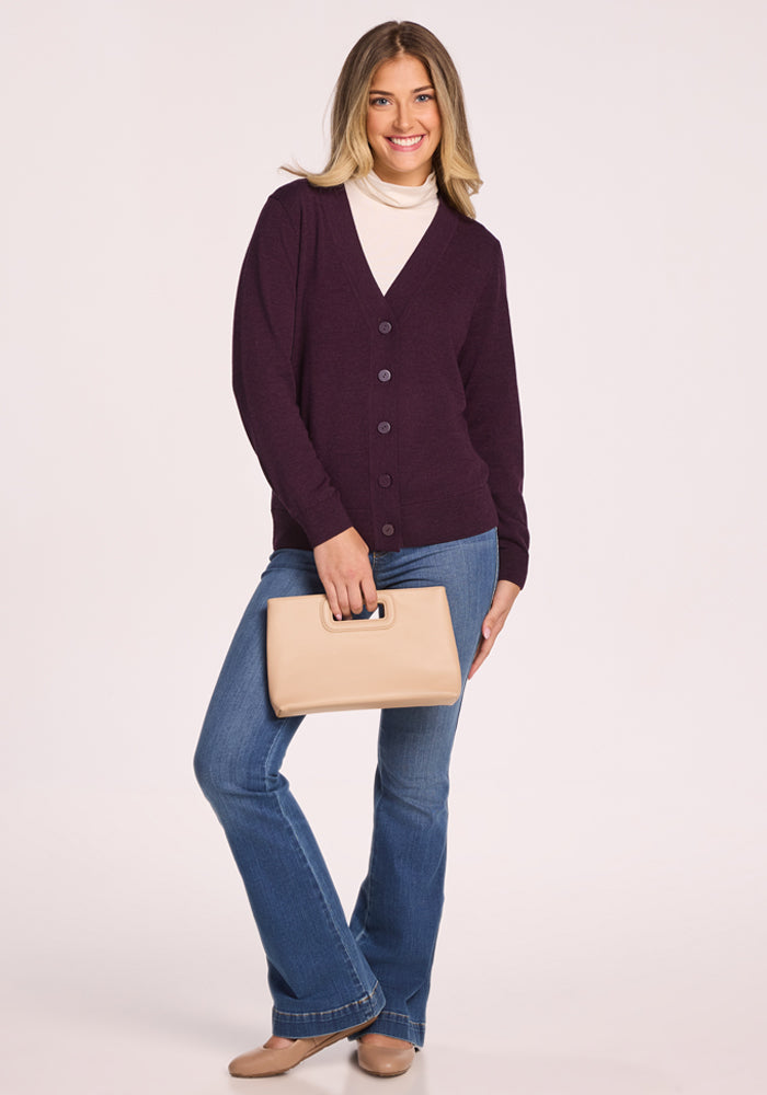 A smiling woman with long blonde hair wears the Woolx Gia Sweater in Deep Plum, paired with a white turtleneck, blue jeans, and beige shoes. She holds a beige handbag and stands against a plain light background.