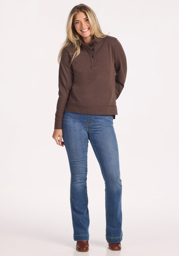 A smiling woman with long blonde hair wears the Woolx Annie 3 Button Pullover in Bracken, paired with blue jeans and brown shoes, standing against a plain light background.