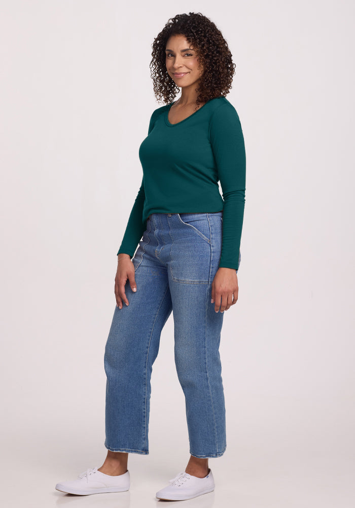 A woman with curly hair stands smiling against a plain background, wearing the Woolx Layla V Neck Top in Riverbank Green, blue jeans, and white sneakers.