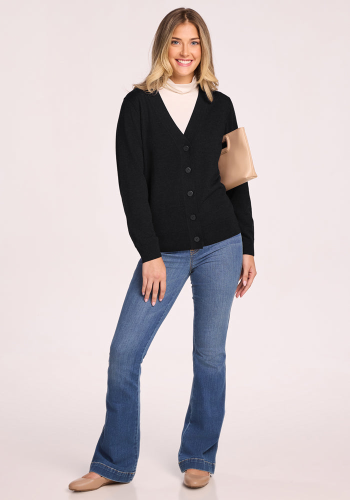 A smiling woman with shoulder-length blonde hair wears the Woolx Gia Sweater in Black, an odor-resistant Merino wool cardigan, paired with a light beige turtleneck, blue flared jeans, beige shoes, and a beige clutch bag against a plain background.