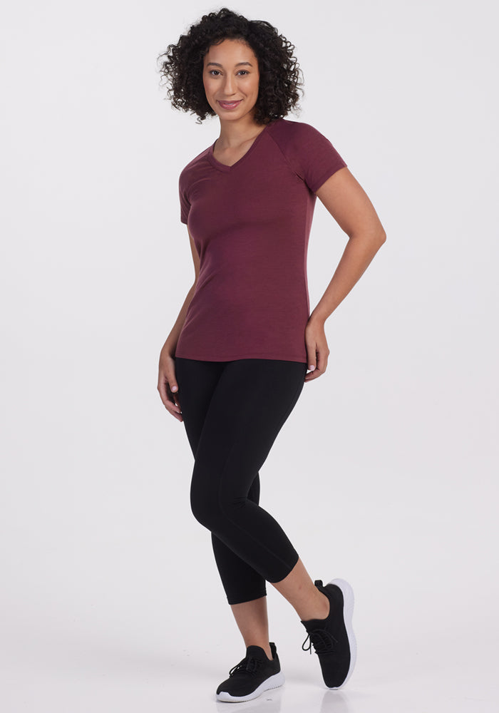A woman with curly hair is standing and smiling, wearing the Woolx Mia V Neck in Grape Harvest, along with black leggings and black athletic shoes, against a plain white background.