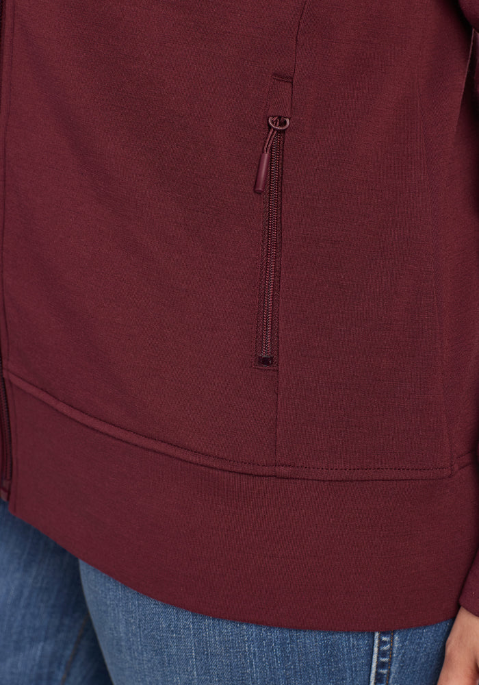 Close-up of a person wearing the Woolx Darby Zip Up Hoodie in Toasted Garnet, featuring a zippered pocket, paired with blue jeans. Only part of the torso and upper leg is visible.