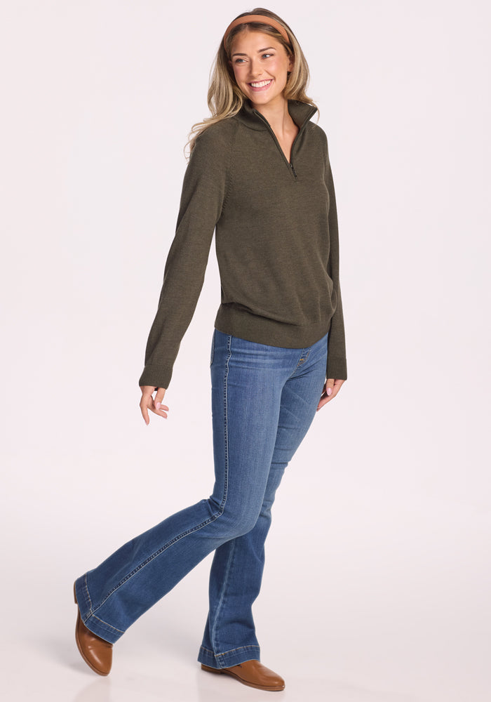 A woman with long, wavy hair wears the Woolx Collins Half Zip Sweater in Pine Needle, blue jeans, and brown shoes. Smiling, she walks with one foot slightly raised against a plain light background.